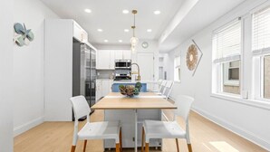 A modern kitchen with white cabinetry, a wooden dining table, and white chairs. - Modern 3BR w/ Backyard, Grill & Outdoor Shower (Ventnor City)