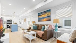 A modern living room with a brown sofa, a coffee table, and a painting of a sunset. - Modern 3BR w/ Backyard, Grill & Outdoor Shower (Ventnor City)
