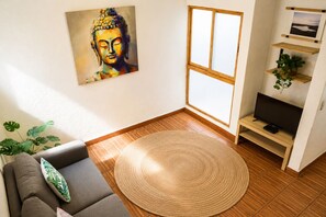 A living room with a sofa, a round woven rug, a television on a wooden stand, a painting on the wall, and a window with a view. - Hikuri House (Tepoztlán)