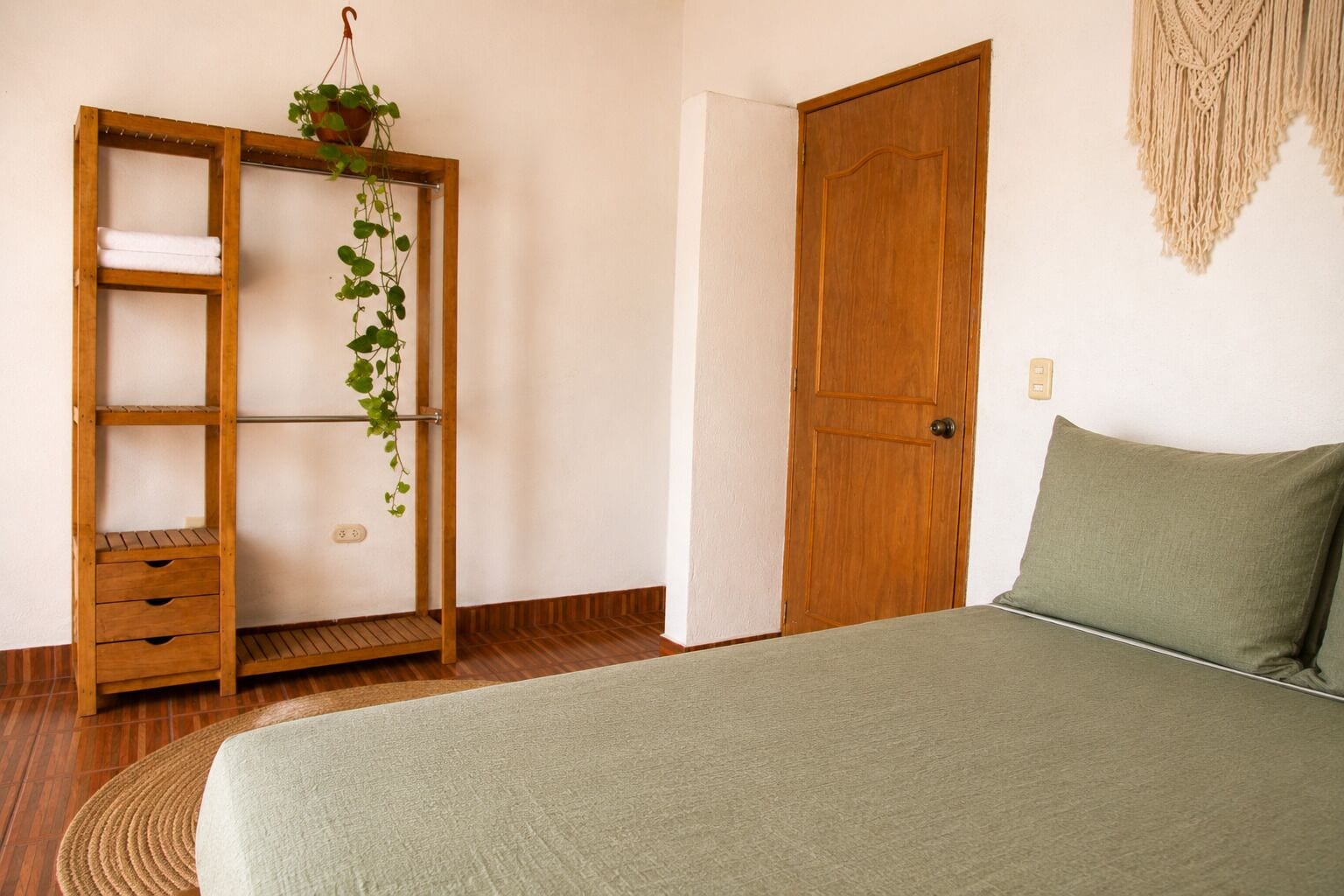 Un dormitorio con una cama, un armario de madera con una planta colgante, una puerta de madera y un macramé en la pared.