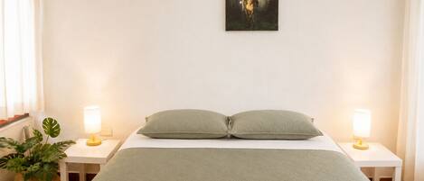 A bedroom with a bed, two bedside tables, a lamp on each side, a wall-mounted painting, and a potted plant.
