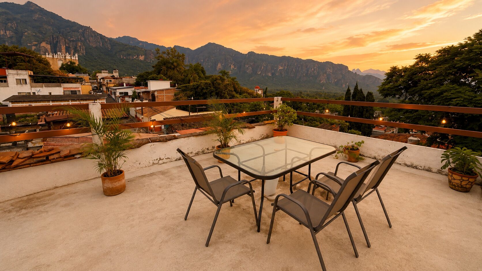 A rooftop terrace with a glass table and chairs, surrounded by potted plants and overlooking a mountainous landscape.