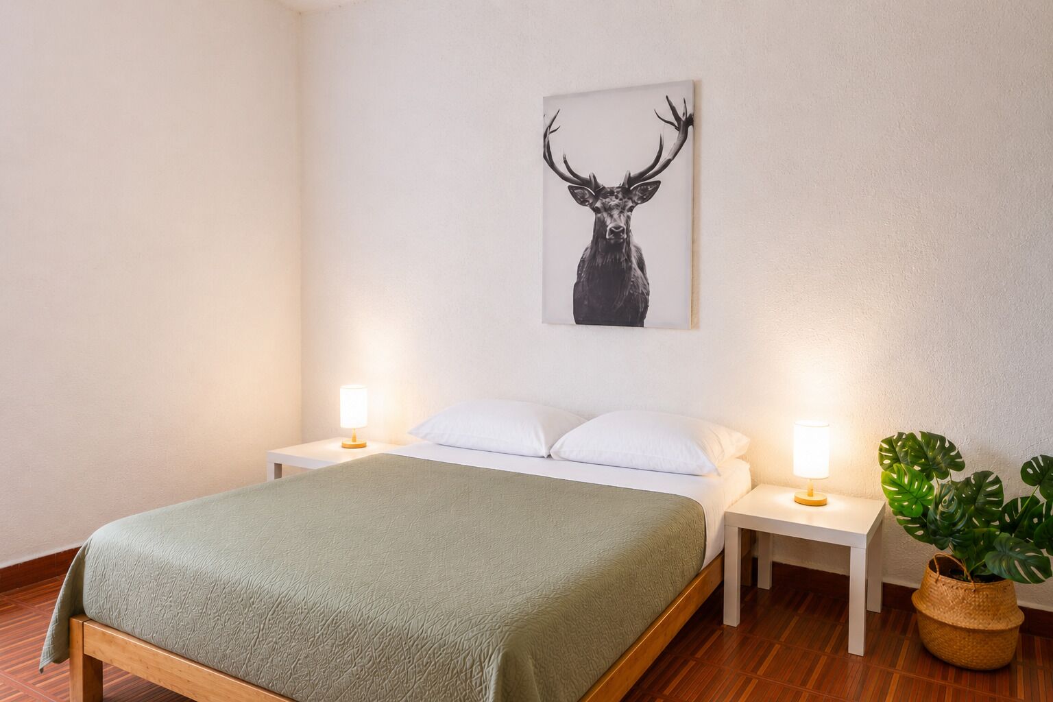 A hotel room with a bed, two bedside tables, a lamp on each table, a wall-mounted picture of a deer, and a potted plant.
