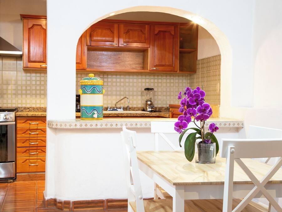 A kitchen with wooden cabinets, a stainless steel range hood, and a tiled backsplash. There is a white dining table with chairs and a potted orchid.