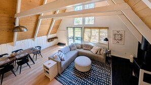 A spacious loft with a high ceiling, wooden beams, a large window, a sofa, a dining table, and a patterned rug. - Harbor Springs Escape, renovated chalet that sleeps 6, near Nubs & Boyne. (Harbor Springs)