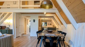 A spacious dining area with a high ceiling and exposed wooden beams, a modern kitchen in the background, and a cozy seating area with a sofa and table. - Harbor Springs Escape, renovated chalet that sleeps 6, near Nubs & Boyne. (Harbor Springs)