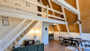 A spacious loft with a high ceiling, exposed wooden beams, a central staircase, a dining area, and a cozy seating area. - Harbor Springs Escape, renovated chalet that sleeps 6, near Nubs & Boyne. (Harbor Springs)