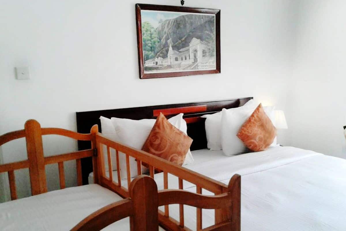 Quarto de hotel com duas cadeiras de madeira, uma cama com roupa de cama branca e almofadas estampadas, e um quadro na parede.