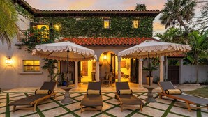 A courtyard with four lounge chairs, a covered seating area, and a building with a tiled roof and ivy-covered walls. - Full Mediterranean Botanical Garden Estate: 2 Adjacent listings, 9bd 5.5bth EV (Sarasota)
