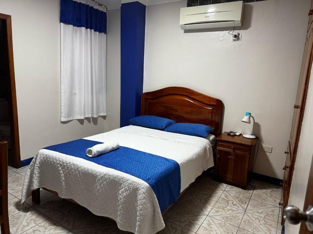 Une chambre avec un lit en bois, une literie blanche et bleue, une table de chevet et un appareil de climatisation fixé au mur.