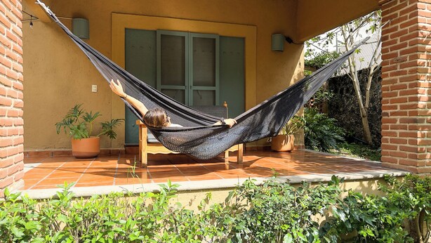 Un hamac sur une terrasse carrelée, avec une chaise en bois, des plantes en pot et un mur en briques.