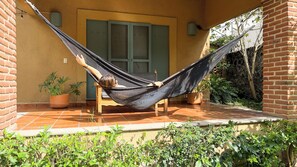 Una hamaca en un patio con baldosas, una silla de madera, plantas en macetas y un muro de ladrillo.