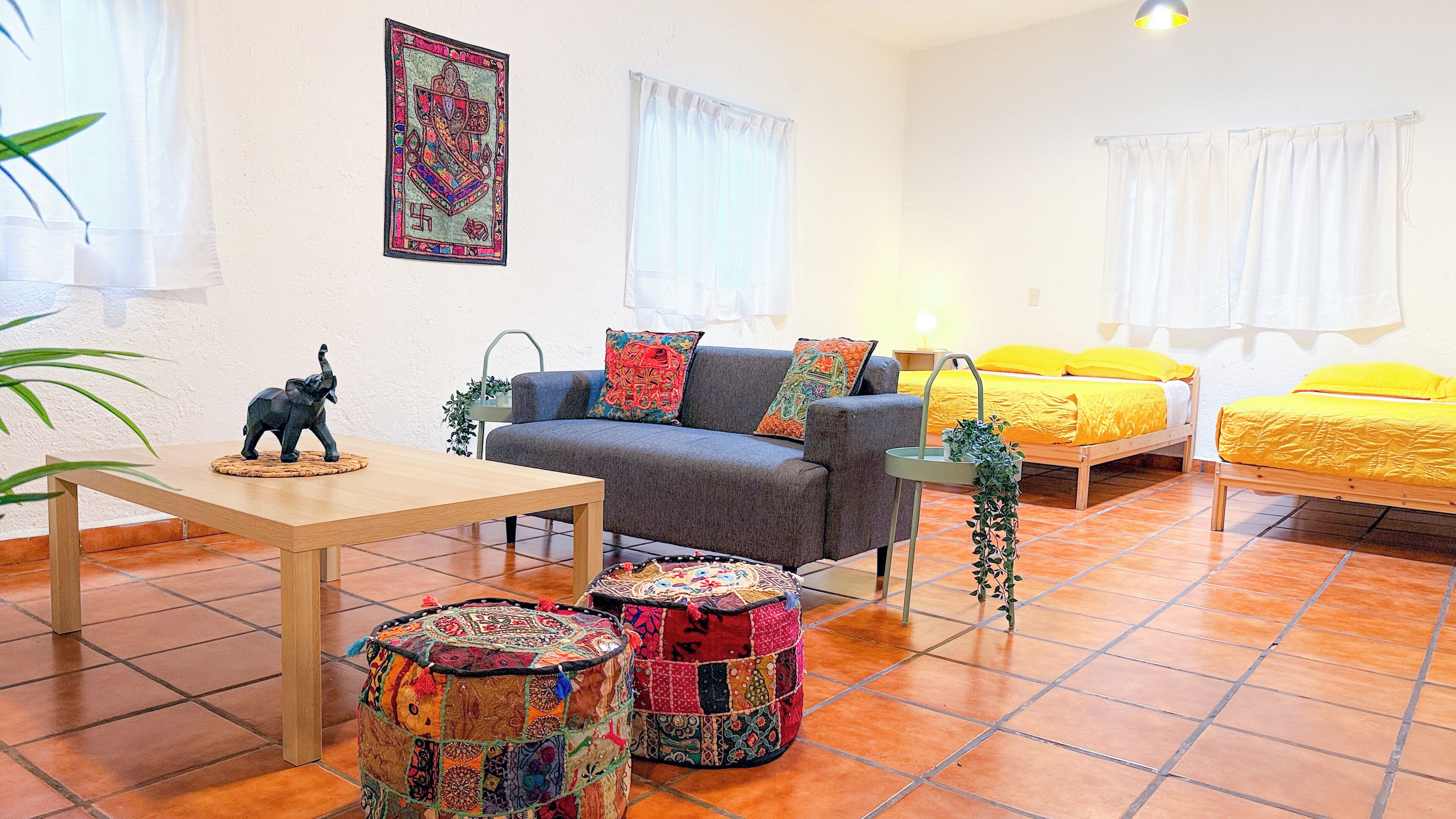 A cozy room with a sofa, a small table, two colorful ottomans, a bed with yellow bedding, and a wall-mounted picture.