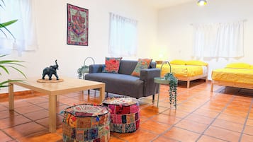 A cozy room with a sofa, a small table, two colorful ottomans, a bed with yellow bedding, and a wall-mounted picture.