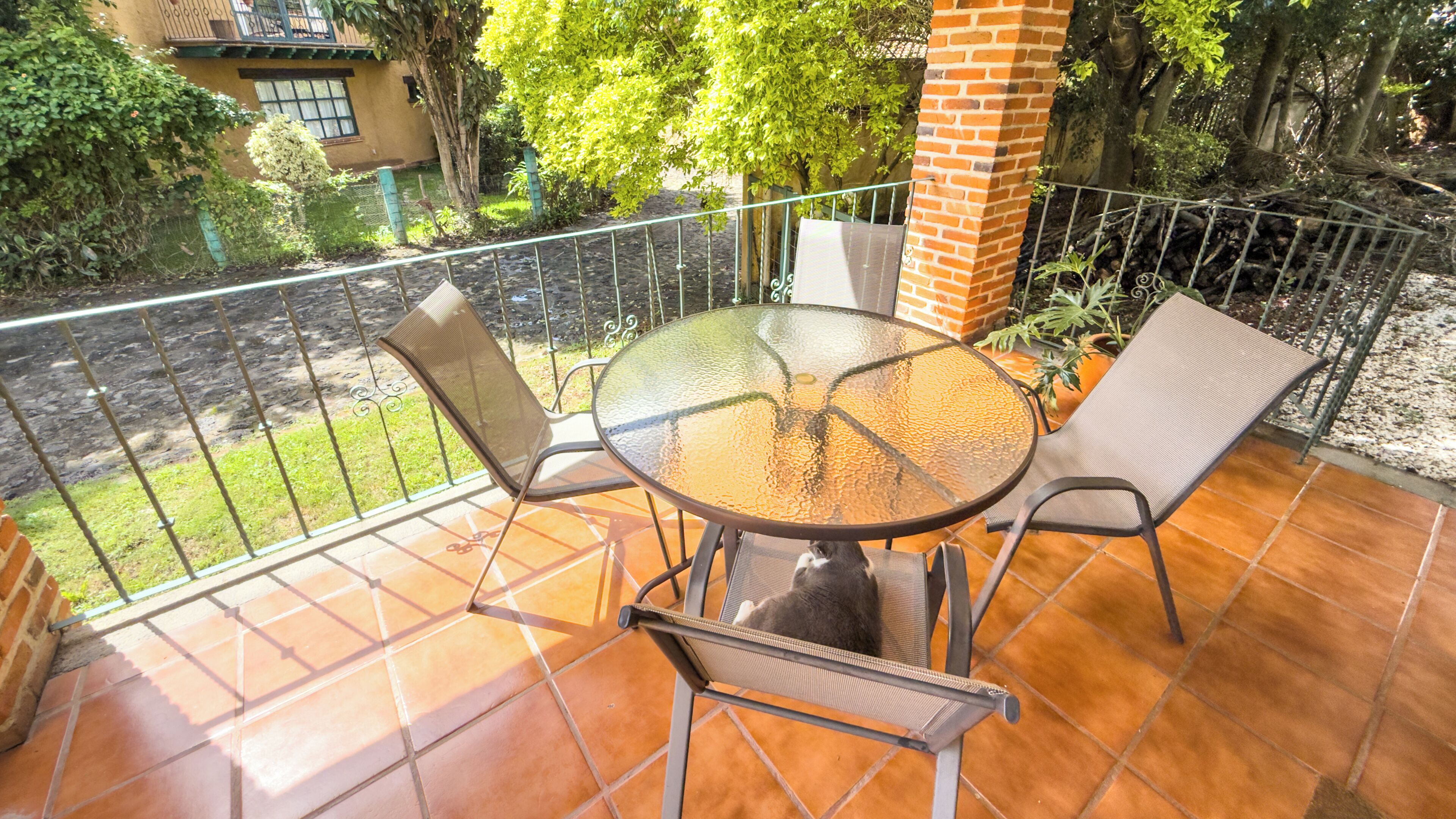 A patio with a round glass table and four chairs, surrounded by a metal fence and a brick pillar.