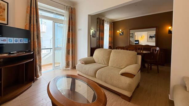 Una sala de estar con un sofá, una mesa de centro de vidrio, un televisor sobre un mueble, un comedor con sillas y una mesa, y un balcón con vistas.