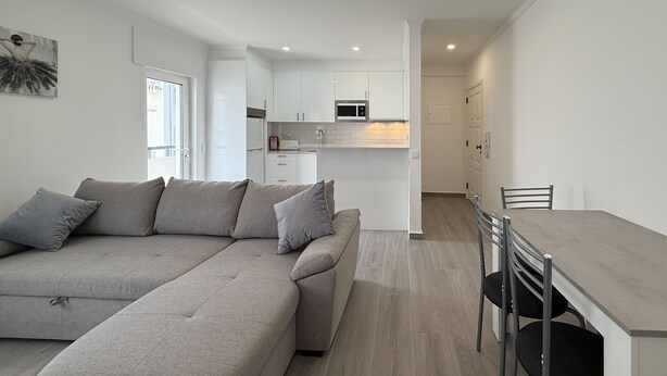 A modern apartment with a grey sectional sofa, a dining area with a table and chairs, and a kitchen with white cabinets and a microwave.