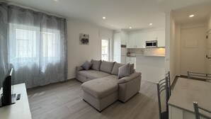 A modern living room with a sectional sofa, a dining area with a table and chairs, and a kitchen with white cabinets and appliances.