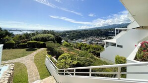 A view from a balcony overlooking a well-maintained garden, a pathway, and a distant cityscape with a body of water and mountains in the background.