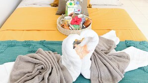 A neatly made bed with a yellow bedspread, a white pillow, and a decorative pillow with a palm tree design. There is a basket with breakfast items and a bottle of water on the bed.