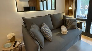 A modern living room with a grey sofa, a large mirror, and a small side table with a remote control and a vase of flowers.