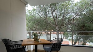A balcony with a wooden table and two black chairs, a potted plant on the table, and a view of trees and buildings.