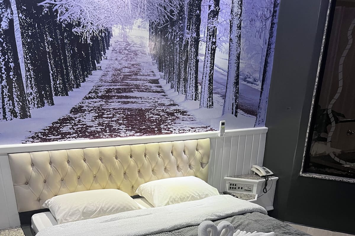 A bedroom with a large bed, a tufted headboard, white linens, and a wall mural of a snowy path.