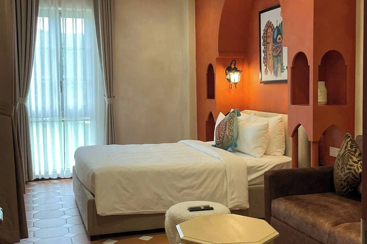 A hotel room with a large bed, a small sofa, a decorative ottoman, and a wall-mounted light fixture.