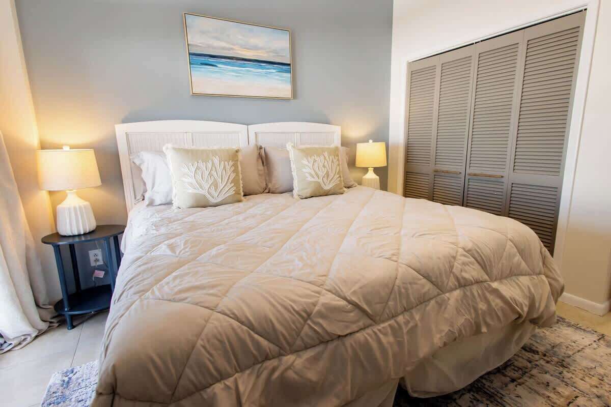 Une chambre avec un grand lit, deux tables de chevet avec des lampes, une commode, un tableau au mur et une armoire avec des portes coulissantes.