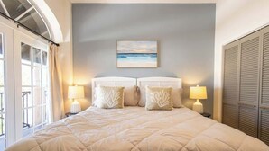 A bedroom with a large bed, two bedside lamps, a painting of a beach, and a window with curtains. - Sandbar Suite | 2 bed near Duval & waterfront (Key West)