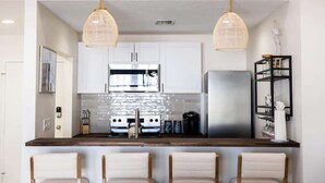 A modern kitchen with white cabinets, a stainless steel refrigerator, and a wooden countertop. There are two hanging pendant lights and a black metal shelf with glassware. - Sandbar Suite | 2 bed near Duval & waterfront (Key West)