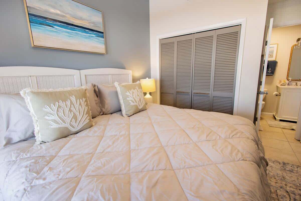 A bedroom with a bed, pillows, a nightstand, a lamp, a closet, and a painting on the wall.