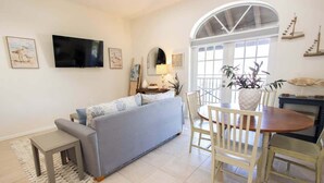 A living room with a grey sofa, a TV mounted on the wall, a dining table with chairs, and a large window with natural light. - Sandbar Suite | 2 bed near Duval & waterfront (Key West)