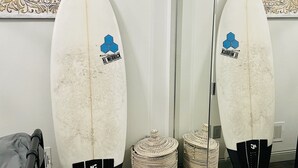 Deux planches de surf appuyées contre un mur orné de panneaux décoratifs, un miroir et des paniers tressés posés sur le sol.
