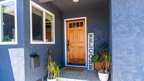 Een blauw huis met een houten deur en een 'WELCOME'-bord. Er staan twee potplanten op de treden.