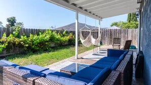 Een overdekt terras met blauwe zittingen, een hangmat en uitzicht op een houten omheining en begroeiing.