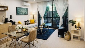 A modern living room with a glass dining table, a sofa with yellow cushions, a coffee machine, and a large window with curtains. - Royal Executive Living Garden Executive (Caulfield North)