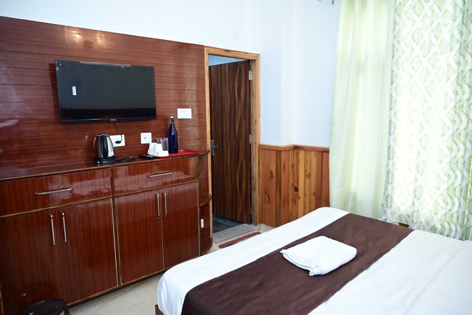 A hotel room with a wooden wardrobe, a flat-screen TV mounted above it, a bed with a brown blanket, and a window with patterned curtains.