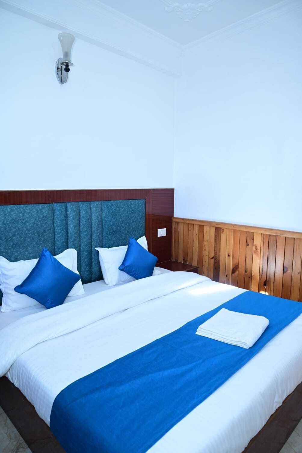 Una cama bien hecha con ropa de cama blanca y azul, un cabecero de madera y una lámpara fijada a la pared.