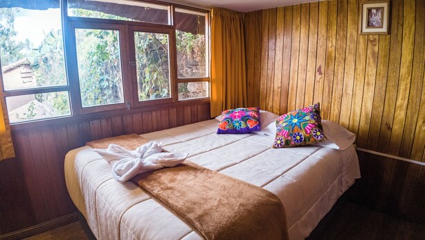 A bedroom with a wooden wall, a bed with a white bedspread, two colorful pillows, and a window with a view of greenery. - CHARMING WOODEN APART IN CENTRAL CUSCO (Cusco)