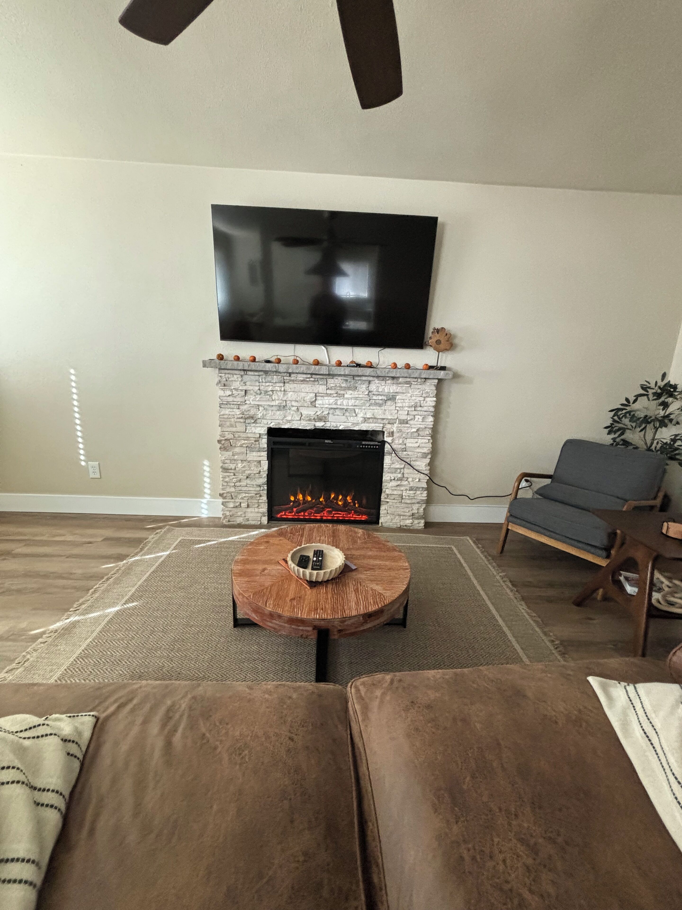 A living room with a stone fireplace, a flat-screen TV mounted above it, a round coffee table with a bowl and remotes, and a brown sofa.