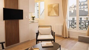Ein modernes Wohnzimmer mit einem Flachbildfernseher, einer Wandpaneele aus Holz, einem schwarzen Sessel, einer Couch, einem Couchtisch mit Tablett und einem Fenster mit Blick auf Gebäude.