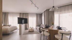 Un salon moderne doté d’un téléviseur à écran plat, d’un canapé, d’une table à manger avec des chaises et d’une grande fenêtre agrémentée de rideaux.