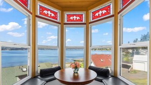 Ein Eckzimmer mit Erkerfenster mit Blick auf einen See, ein runder Tisch mit Blumenvase und schwarze Ledersitzmöbel.