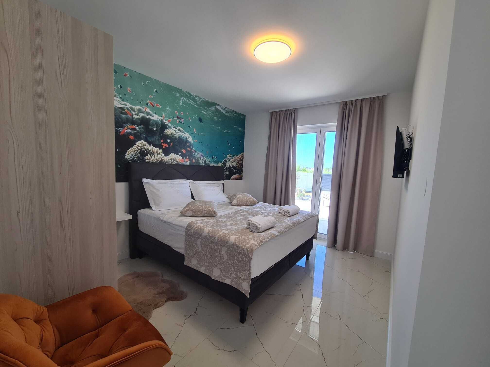 A hotel room with a bed, a large mural of an underwater scene, a ceiling light, and a view of the outdoors through a sliding door.