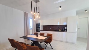A modern dining area with a rectangular table and six chairs, a kitchen with white cabinets, and a door in the background. - New villa Angelo 2025 ( family and pet friendly) (Privlaka)