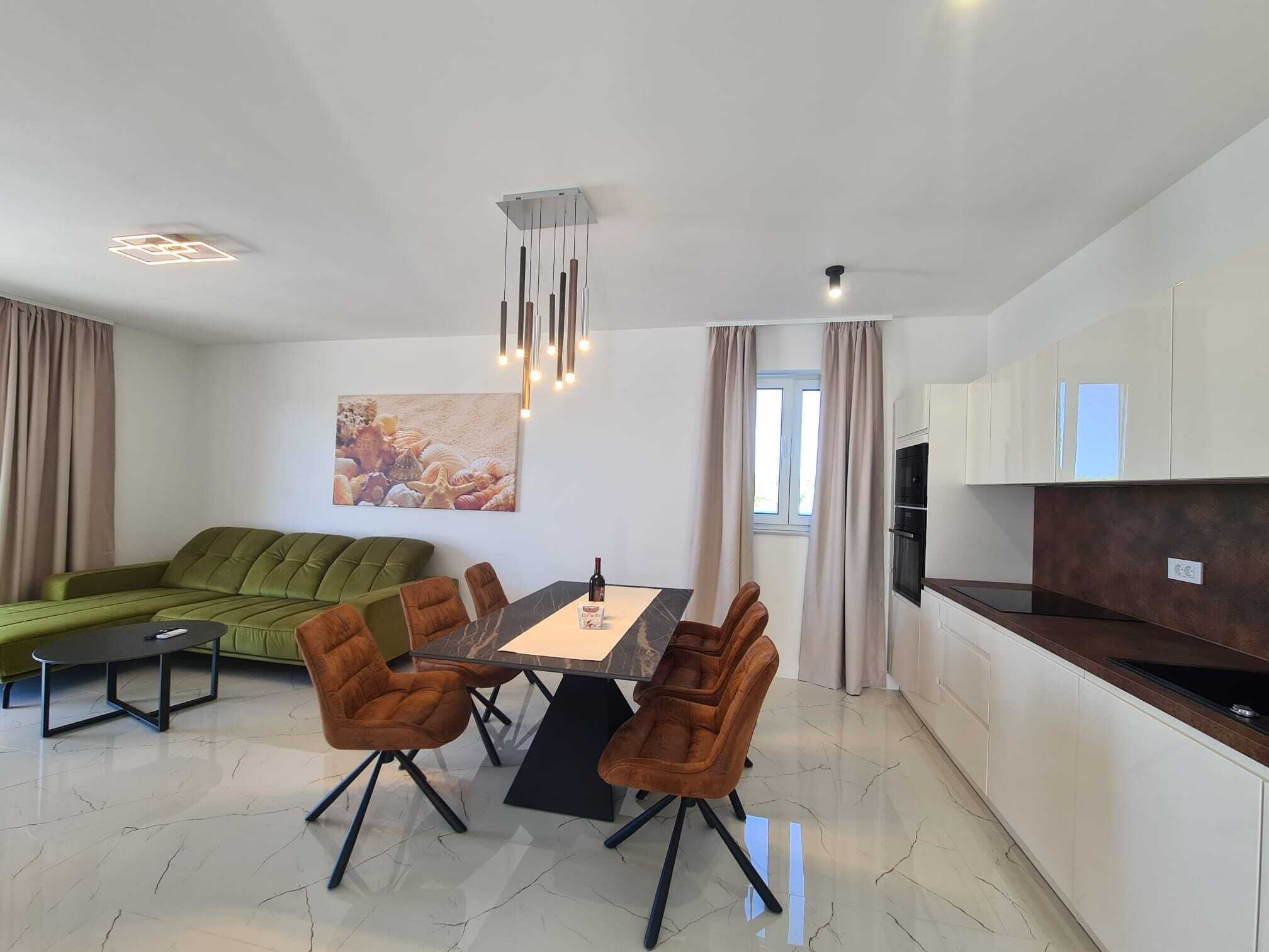 Uma sala de estar moderna com um sofá verde, uma mesa de jantar com quatro cadeiras, e uma área de cozinha com armários brancos e bancada escura.