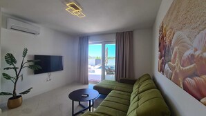 A modern living room with a green sofa, a flat-screen TV, a potted plant, and a large painting of seashells. - New villa Angelo 2025 ( family and pet friendly) (Privlaka)