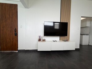 Un salon moderne avec un téléviseur à écran plat fixé sur un meuble de rangement blanc, une porte en bois et un coin cuisine à l’arrière-plan.