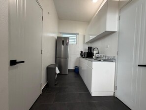 A narrow hallway leading to a kitchen with white cabinets, a stainless steel refrigerator, and a small trash bin.